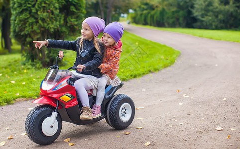 坐在绿色公园的玩具摩托车上坐着的小可爱姐妹自行车游戏快乐喜悦孩子乐趣婴儿运输活动女孩背景图片