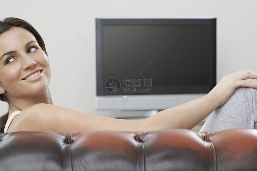 在沙发上当着平板屏幕电视的女青年娱乐活动成年黑发客厅摄影真皮成人女士微笑图片