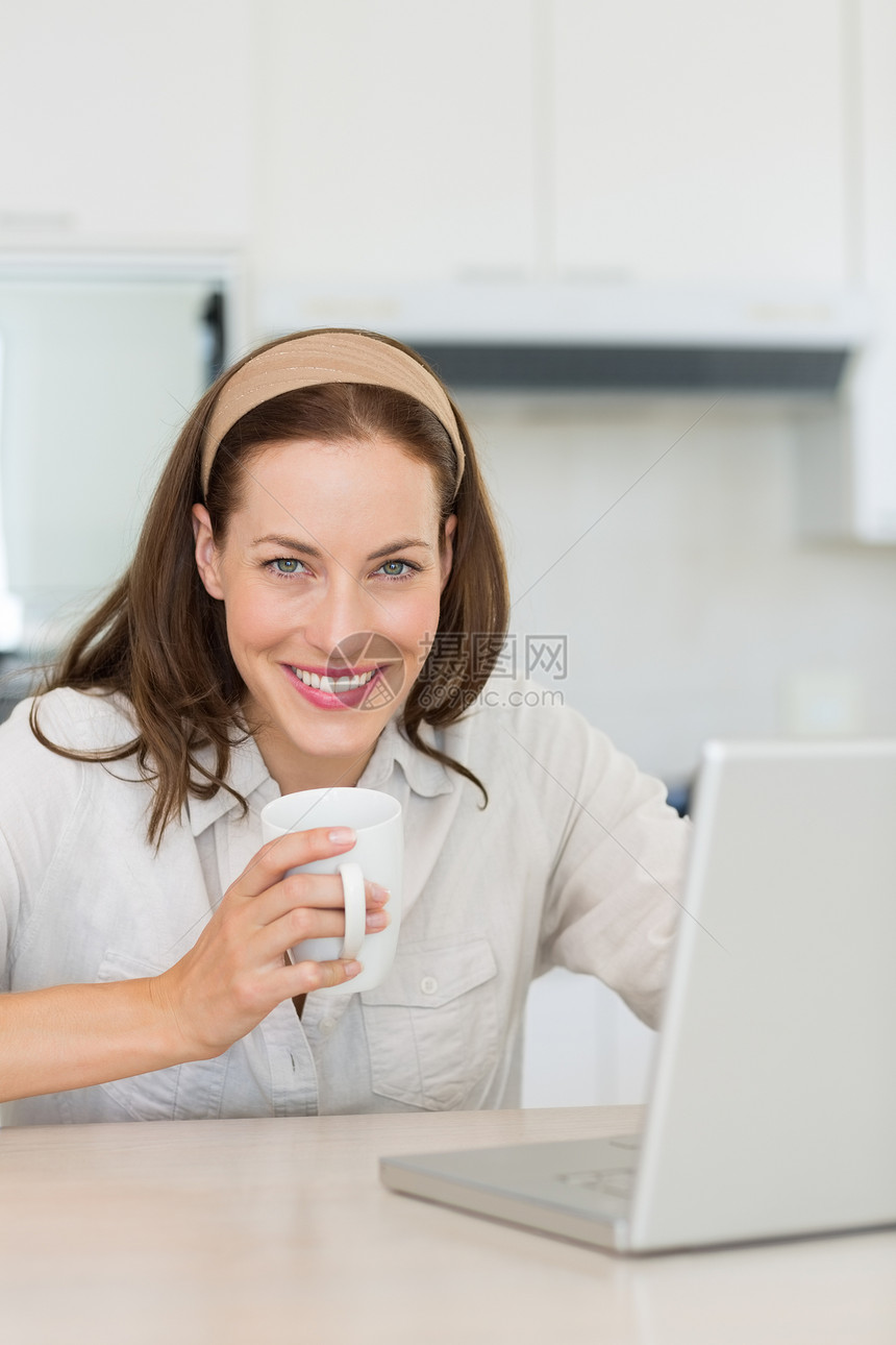 带咖啡杯和厨房笔记本电脑的笑女人棕色咖啡杯子女性闲暇柜台女士房子头发技术图片