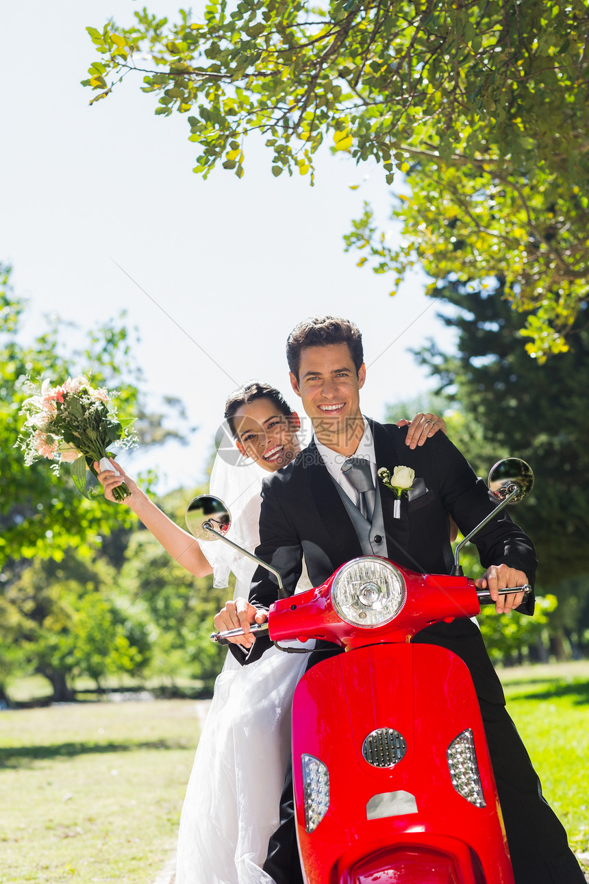 新婚夫妇坐在公园的摩托车上乐趣新郎已婚运输妻子男性丈夫花束树木婚纱图片
