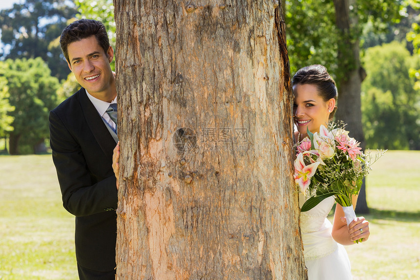 在公园树干后面的一对新婚夫妇图片