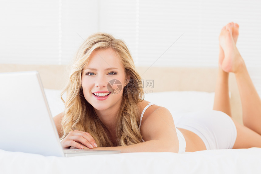 用笔记本电脑在床上微笑着年轻金发美女图片