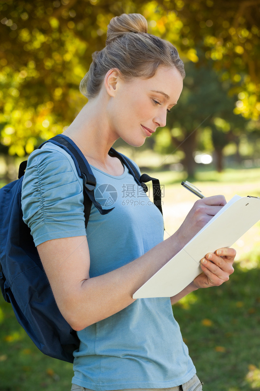 美丽的年轻女子在公园剪贴板上写作旅行绿地草地树木头发笔记专注背包美好生活女士图片