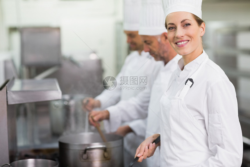 快乐的厨师在炉灶做饭时对着照相机微笑滚刀职员厨艺酒店烹饪女士炉顶团队制服合作图片