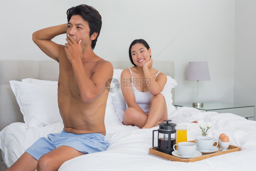 微笑的女人看着她的男朋友打哈欠和伸展公寓咖啡卧室女性咖啡馆被子男性羽绒被家庭快乐图片