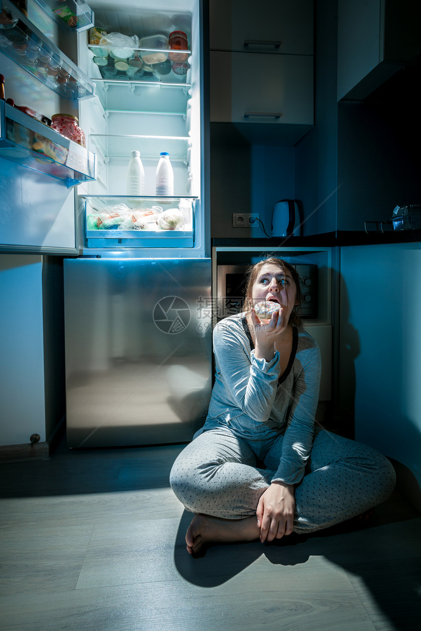 穿着睡衣的女人晚上在冰箱旁边的地板上吃饭图片