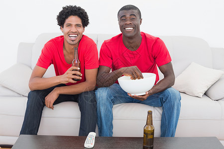 红红色足球球迷坐在沙发上 啤酒和爆米花服装休闲亲密感活力友谊遥控家庭生活观众闲暇男性背景图片