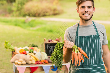 手持胡萝卜的英俊农民桌子蔬菜农贸市场微笑快乐黑板乡村围裙闲暇失速背景图片