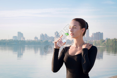 年轻女子在清晨的河水和城市背面喝水背景图片