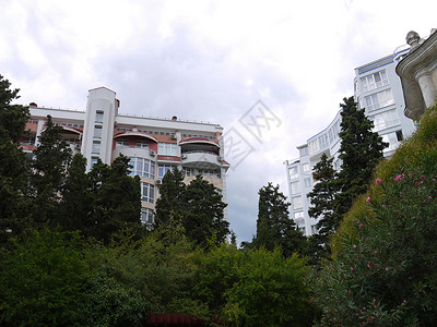 林线旅馆绿树和开花林后面的绿树和鲜花灌丛 在云天背景下高亮的旅馆大楼或现代公寓楼背景