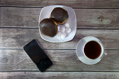 一杯红茶放在碟子上 盘子里放着两个涂有巧克力糖衣的甜甜圈和大块面包 放在智能手机旁边的木桌墙上 咖啡时间 休闲时光 特写杯子厨房背景图片