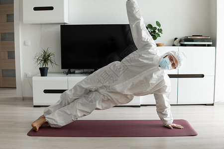戴着医用口罩和防护服的年轻人正在家里的垫子上练习瑜伽 在 COVID19 检疫概念期间待在家里 在家在线瑜伽 冠状病毒大流行 健背景