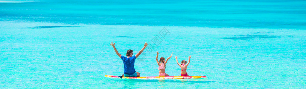 蓝色女孩暑假期间在冲浪板上的小女孩和年青父亲喜悦海洋热带海浪幸福冲浪家庭冒充泳装女孩背景