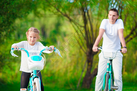 骑自行车的父亲年幼的父亲和小女孩在夏暖日骑自行车公园家庭孩子农村头盔女孩乐趣姐姐娱乐父母背景