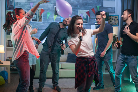 在派对上唱卡拉OK的 兴奋的年轻女子歌曲唱歌伙计们气球酒精音乐麦克风沙发乐趣朋友们背景图片