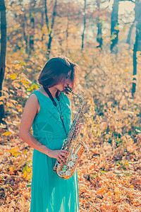 次中音号穿着长蓝色裙子的深褐色女孩 在秋森林里演黄色萨克斯风剧背景