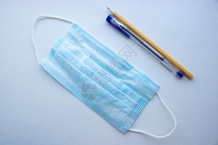 防护层透明膜白色背景的钢笔和铅笔旁边放着一个带橡胶耳带的外科或医用口罩 典型的 3 层医生口罩 用于遮盖口鼻 防护概念背景