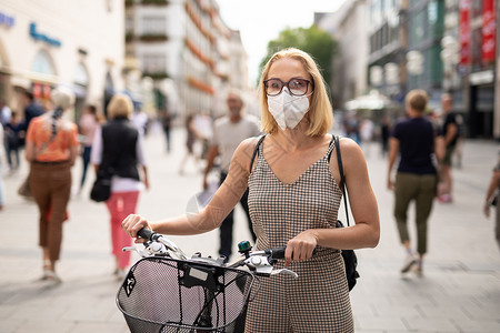 妇女用自行车在行人城市街道上走路 在公共场合戴医疗面罩 以防止科罗纳病毒传播 在贪食流行期间是新常态流感症状男人女孩太阳镜口罩面背景图片