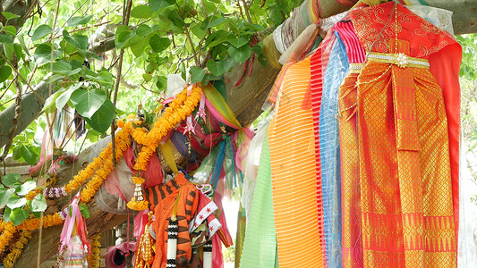 五颜六色的供服在祭坛附近的树上 万物有灵祭坛附近树枝上挂着一堆鲜艳的传统服装 作为送给泰国烈酒的礼物背景
