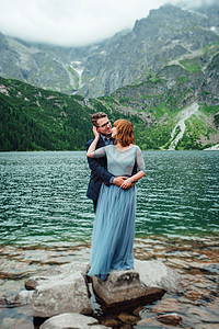 年轻一对夫妇在环绕山峰的湖边散步新娘公园支撑天空成人花岗岩石头婚礼后台会议背景图片