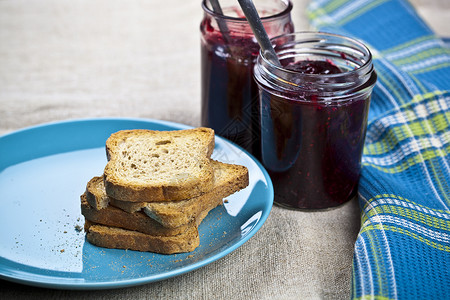 蓝色罐子蓝陶瓷板上的谷物面包切片 罐子和勺子中的自制樱桃和野莓果酱 以及生锈木桌底的餐巾纸背景