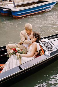 新郎装在威尼斯的意大利婚礼上 新娘和新郎骑着经典的木制歌多拉 沿着狭窄的威尼斯运河走目的地花束金发家庭蜜月奢华棕色玫瑰丈夫新婚背景