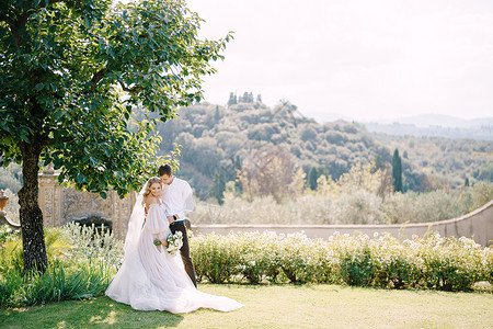 真正的婚礼在意大利佛罗伦萨的一个古老的别墅酒庄举行婚礼 新娘和新郎在树荫下 新婚夫妇在花园里散步羽毛投标拥抱金发女郎薄纱白玫瑰丝带花朵婚纱背景