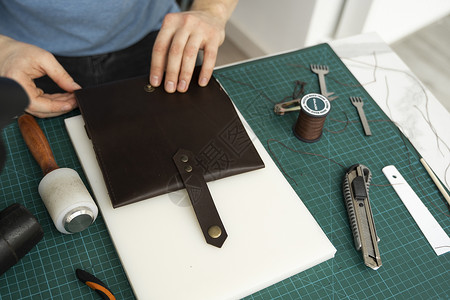 男士手在他的工作室里制作皮革钱包 与棕色天然皮革的工作过程 拿着制作工具的工匠剪刀前锋机器作坊皮肤制造业做工锥子维修鞋匠背景图片