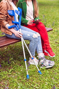 两名妇女 一名健康 一名脚扭伤 坐在长凳上拐杖公园时间腿部女性闺蜜闲暇草地长椅朋友们背景图片