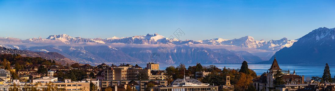 以山为背景的建筑洛桑市风景与日内瓦湖(Lake Leman)的天空全景以及雪将法国阿尔卑斯山在阳光明媚的秋天以蓝云为背景汇集在一起 瑞士沃州Va背景