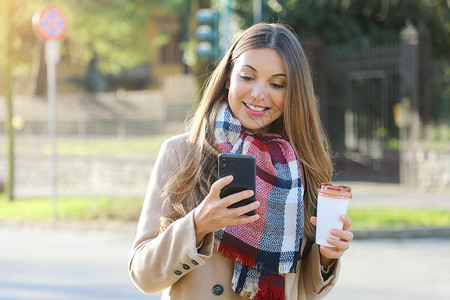 来电话了穿着大衣和围巾的年轻快乐女性在城市街道上用手机使用她的电话 饮酒夺走了咖啡背景