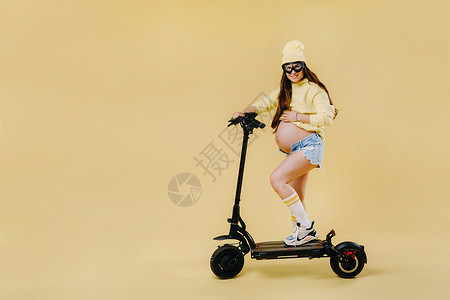 一名身着黄色衣服的怀孕女孩 她坐在一辆离世黄色背景的电动摩托车上青年骑术运输生态帽子车辆女士笑声成人夹克背景