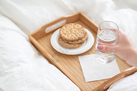 女性手在托盘上举着水杯 床上装着饼干食物节食白色营养饮食玻璃桌子背景图片