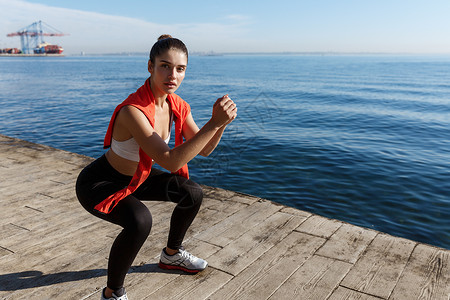 外出打工素材外出拍摄有吸引力的健身女在海上附近打工 蹲下锻炼和看照相机女孩海洋活动训练码头下蹲有氧运动女性成人跑步背景