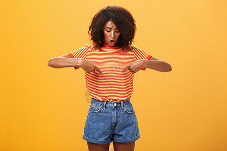 我头和胃很疼忧心忡忡 质疑和惊讶的非洲裔美国女性的画像 头发卷曲 看着并指着胃或腹部 在橙色背景下感到不适和健康问题学生广告成人工作室橙子爆背景
