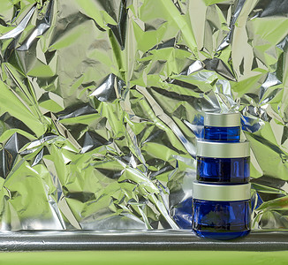 带灰色塑料盖的蓝色玻璃罐 用于化妆品 背景为皱巴巴的铝箔 化妆品霜 面膜 血清品牌小样推广塑料凝胶包装治疗润肤产品玻璃卫生背景图片