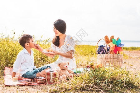 零食酷站欣赏美丽的亚洲妈妈在野餐时在草地上给儿子喂零食 母亲和儿子一起玩耍 在母亲节庆祝和欣赏概念 夏天的人和生活方式的主题背景