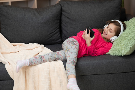 短句心情记录戴着无线耳机的快乐小女孩在沙发上大笑 使用移动在线播放器应用拿着手机听着最喜欢的音乐 在家里戴着耳机享受宁静的心情背景