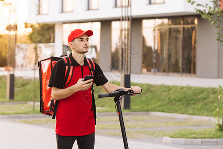 红色背包男性送信员用红色热背包在街上行走 配有电动摩托车使用智能手机导航 产货员雇员在日落时提供在线订单客户服务背景