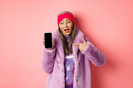 在线购物和时尚概念 酷的亚洲奶奶穿着时装假毛皮大衣 将手指左手放在空白智能手机屏幕上 检查互联网宣传 粉红色背景标识女性广告技术背景图片