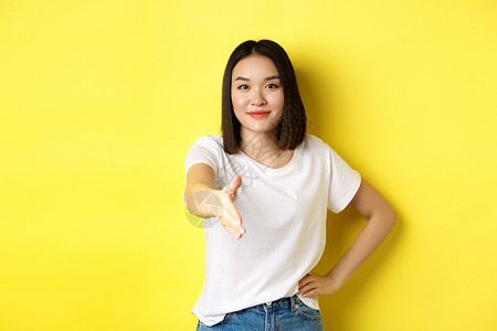 自信的亚洲女性穿着白色 T 恤 伸出手握手和打招呼 打招呼 自我介绍 站在黄色背景上手势快乐情感广告工作室成人黑发青少年发型女孩背景图片