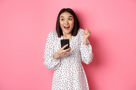 在线购物和美容理念 兴奋的亚洲女性在互联网上获胜 手持智能手机欢欣鼓舞 微笑着开心庆祝 站在粉红色背景中互联网店铺电话黑发技术网背景图片