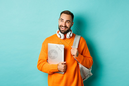 背着背包和耳机的快乐男人 拿着笔记本电脑 微笑着 左顾右盼 站在绿松石背景上胡子自由职业者情感毛衣横幅胡须男性工作标识黑发背景图片