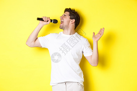 麦克风促销主图年轻男子歌手拿着麦克风 达到高音调和唱卡拉OK 站在黄色背景上胡须闲暇商业成人手势微笑促销卡拉ok潮人广告背景