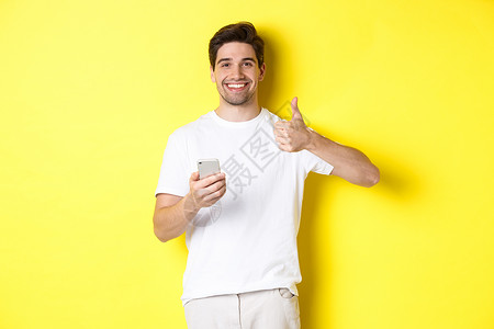 快乐满意的男人拿着智能手机 竖起大拇指表示赞同 在网上推荐一些东西 站在黄色背景上男性微笑广告手势电话黑发商业工具闲暇成人背景图片