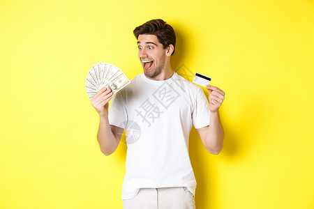 快乐的人看着钱 拿着信用卡 银行信贷和贷款的概念 站在黄色背景上情绪购物闲暇金融微笑手势男性潮人成人工作室背景