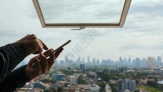 在打开的窗户和雅加达市全景的背景下 一名男子在智能手机上键入一条消息 双手合十背景图片