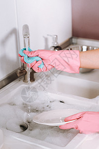 手托盘子戴粉红色手套的家庭主妇女孩用洗涤剂在水槽里用手洗碗 女孩在家里打扫房子 戴着手套洗碗盘子洗衣机脸盆清洁度妻子餐具菜肴女士海绵眼镜背景
