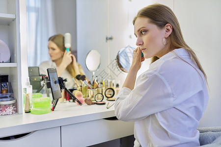 化妆课年轻女士坐在化妆桌前 照着镜子做化妆和发型魅力女孩视频博主电话刷子头发记录化妆台手机背景