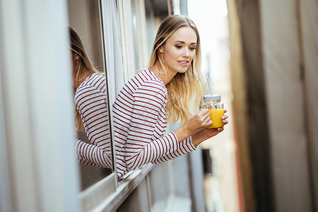 年轻女子喝着一杯天然橙汁 从家窗外倾斜而下 从家里走出来食物玻璃稻草成人饮食女性窗户头发橙子水果女孩高清图片素材